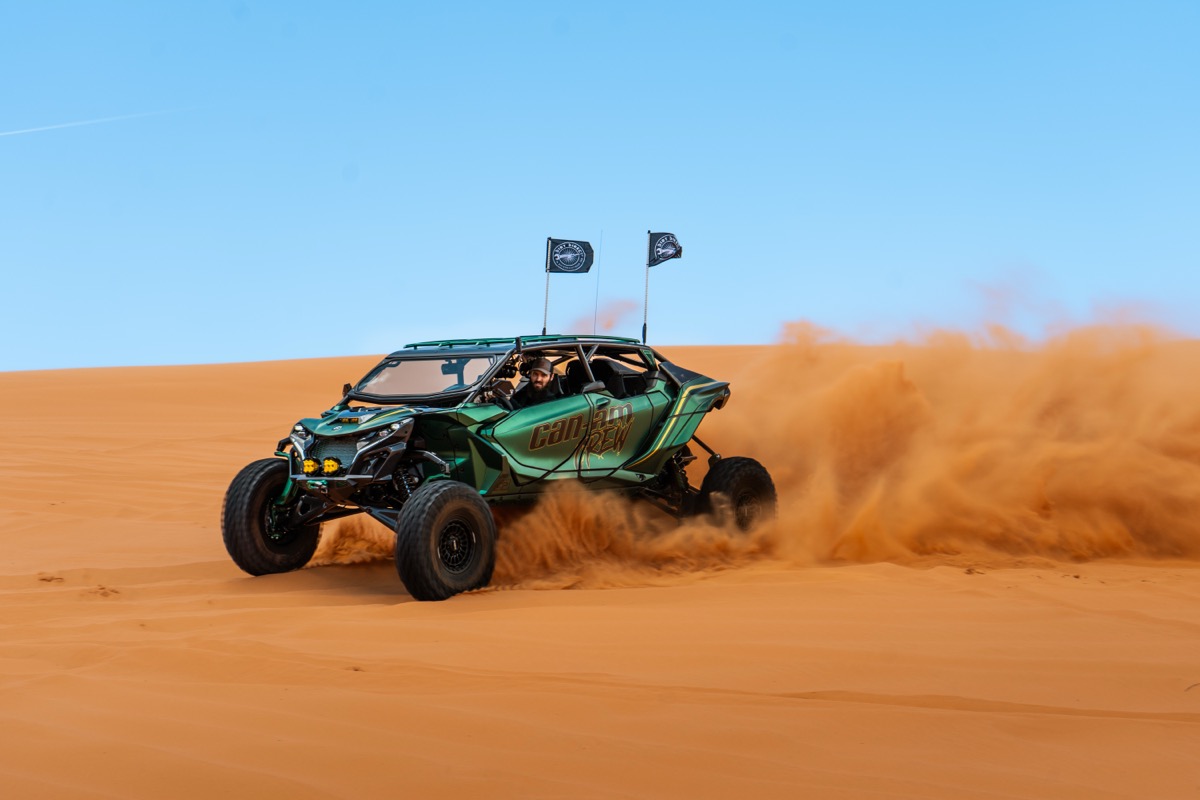 Can-Am Maverick X3 with Can-Am Crew branding throwing a massive sand roost while carving across orange dunes at Sand Mountain Utah