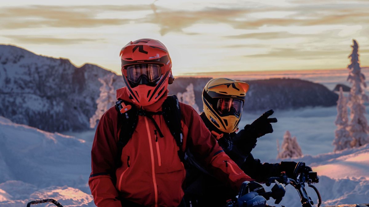 Two snowmobile riders in full gear on a snowy mountain at golden hour with snow-covered trees and mountain views